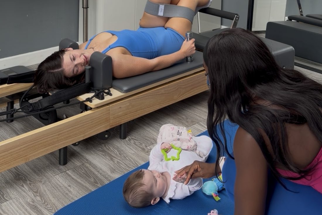 A mum training on the reformer during a postnatal session, her baby on the mat beside her with the coach close by